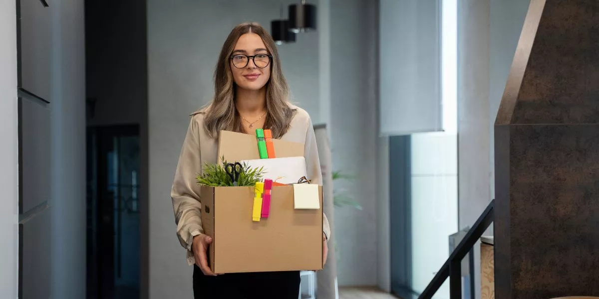 Mujer saliendo de una oficina con una caja de pertenencias tras presentar una renuncia voluntaria de trabajo.