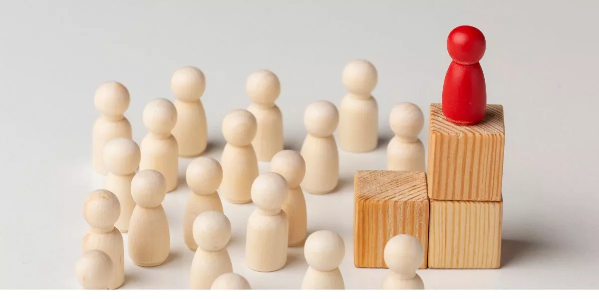 Figura roja destacada sobre bloques de madera frente a un grupo de figuras claras, representación visual de los tipos de liderazgo empresarial y la influencia del líder en el equipo.