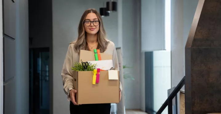 Mujer saliendo de una oficina con una caja de pertenencias tras presentar una renuncia voluntaria de trabajo.