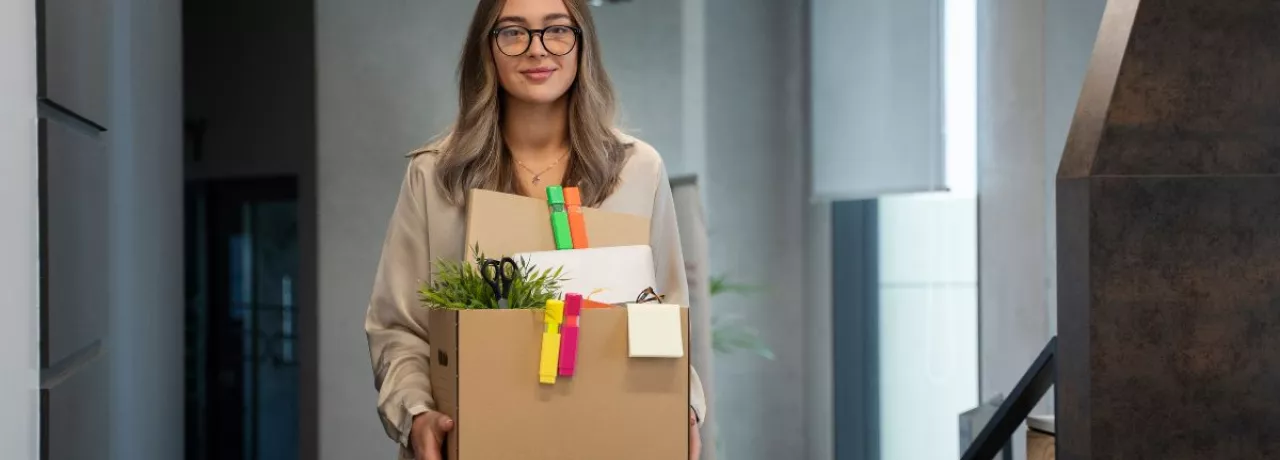 Mujer saliendo de una oficina con una caja de pertenencias tras presentar una renuncia voluntaria de trabajo.