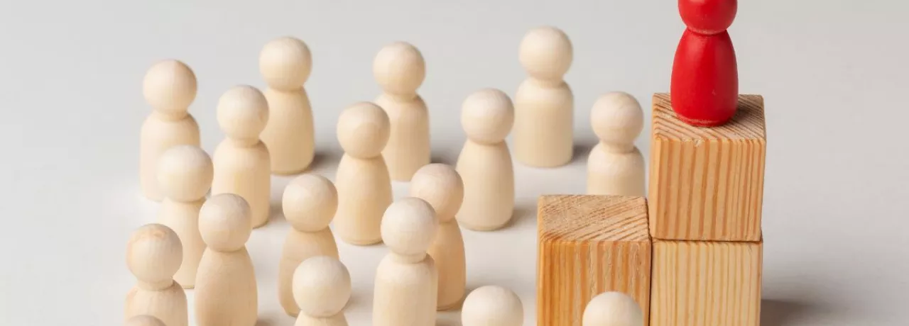 Figura roja destacada sobre bloques de madera frente a un grupo de figuras claras, representación visual de los tipos de liderazgo empresarial y la influencia del líder en el equipo.