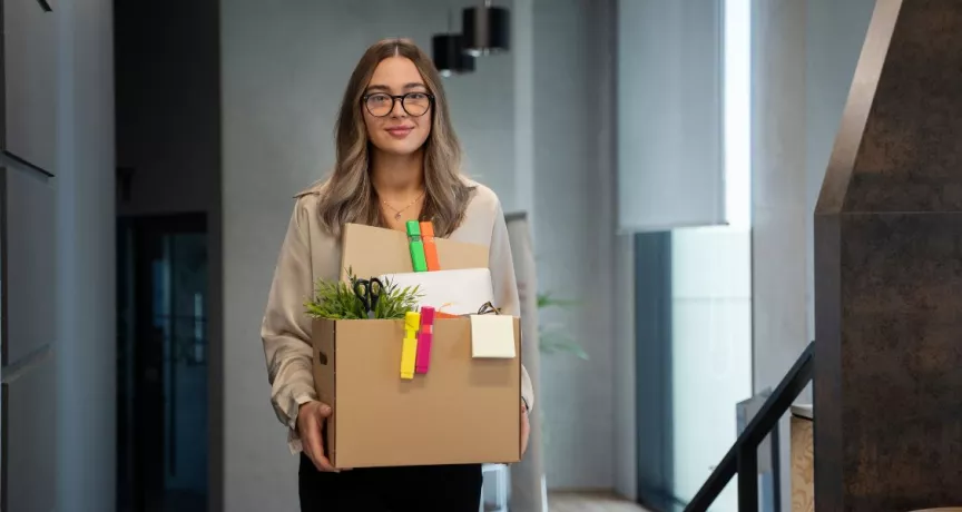 Mujer saliendo de una oficina con una caja de pertenencias tras presentar una renuncia voluntaria de trabajo.
