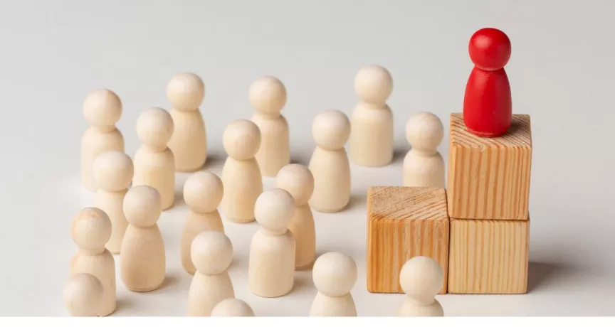 Figura roja destacada sobre bloques de madera frente a un grupo de figuras claras, representación visual de los tipos de liderazgo empresarial y la influencia del líder en el equipo.