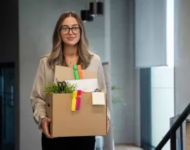 Mujer saliendo de una oficina con una caja de pertenencias tras presentar una renuncia voluntaria de trabajo.