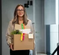 Mujer saliendo de una oficina con una caja de pertenencias tras presentar una renuncia voluntaria de trabajo.