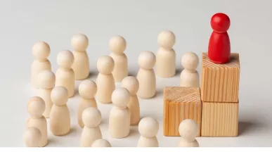 Figura roja destacada sobre bloques de madera frente a un grupo de figuras claras, representación visual de los tipos de liderazgo empresarial y la influencia del líder en el equipo.